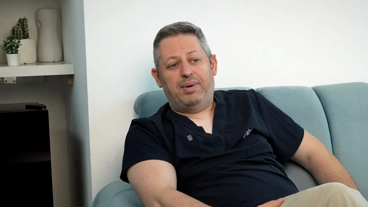 ד״ר חגלי בסרטון על השתלות שיניים בחו״ל, יושב על ספה במרפאה ומסביר למה חשוב מעקב קרוב לבית